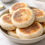 Homemade sourdough English muffins, golden brown and fluffy, on a kitchen table.